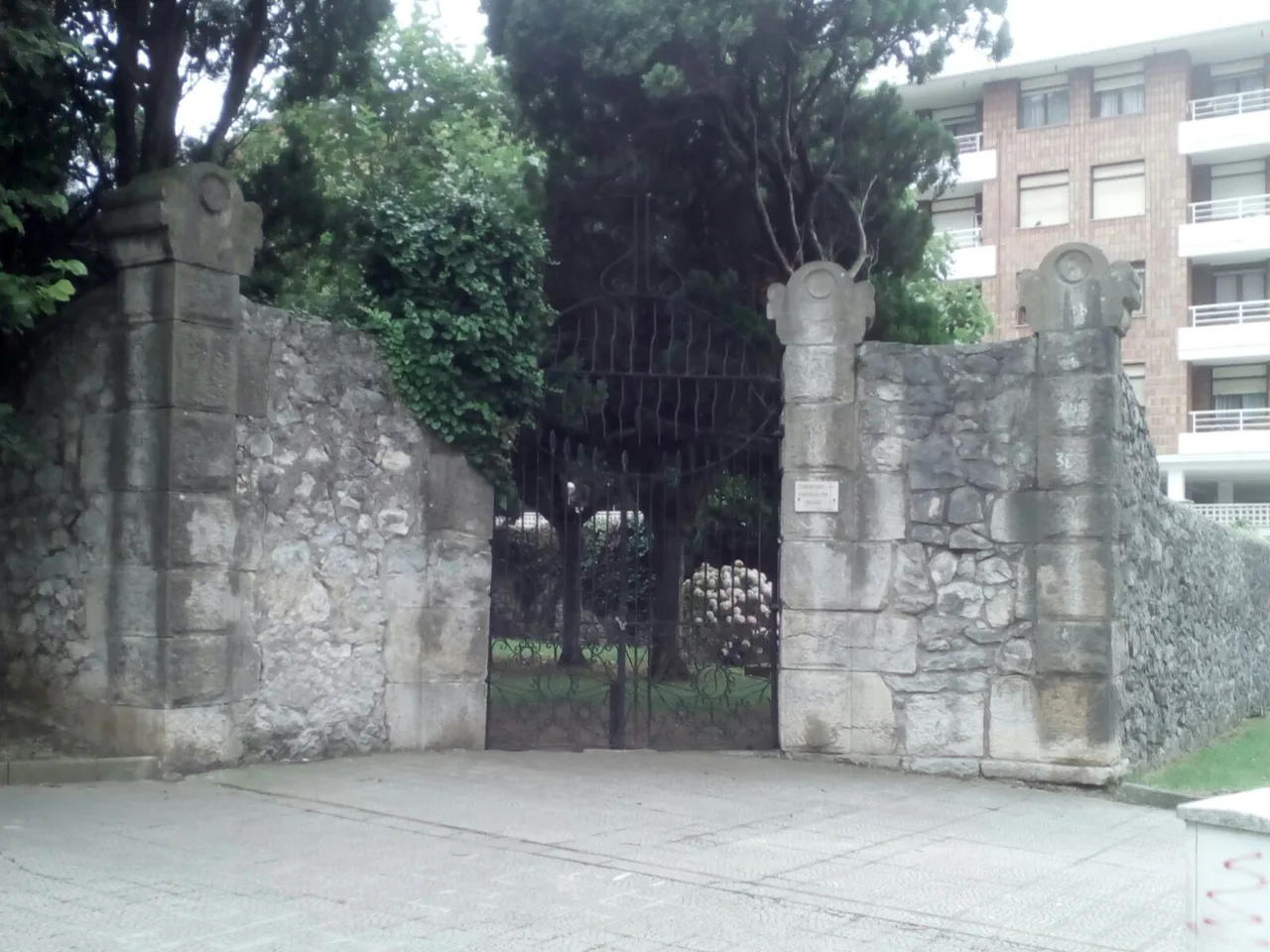 Portada de piedra y puerta de hierro del cementerio protestante de Cazoña, con árboles tras la verja y viviendas al fondo
