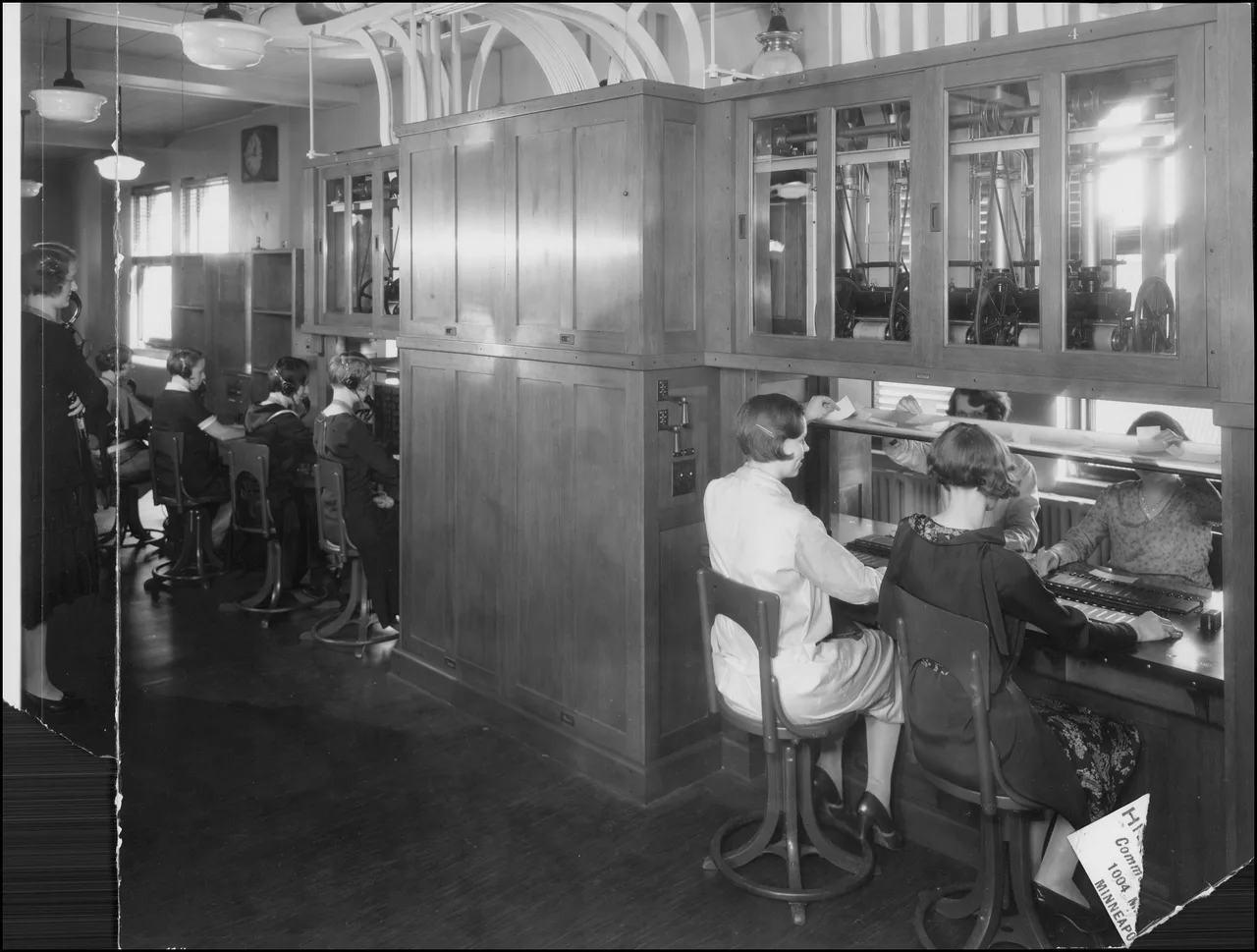Varias mujeres trabajan con auriculares entre escritorios y un cuadro telefónico en una central de 1930