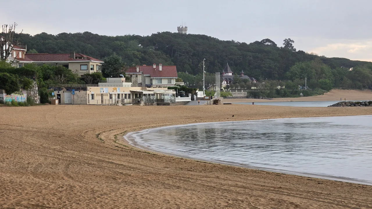 Curva de arena y orilla de la playa de la Magdalena, con el balneario y la península al fondo